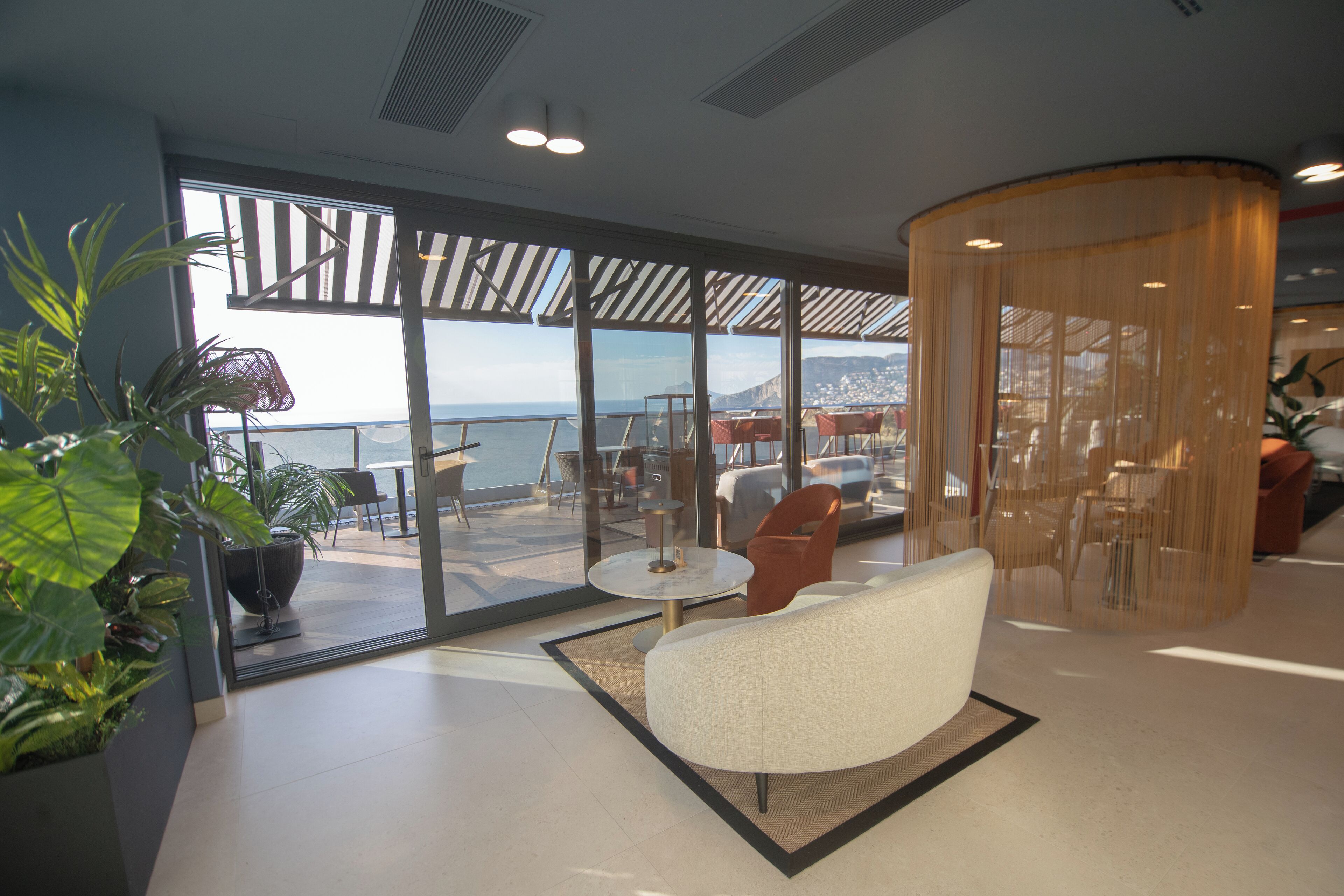 Eine moderne Hotelhalle mit einer großen Glaswand, die Meerblick bietet, und einem Balkon mit Außenmöbeln.