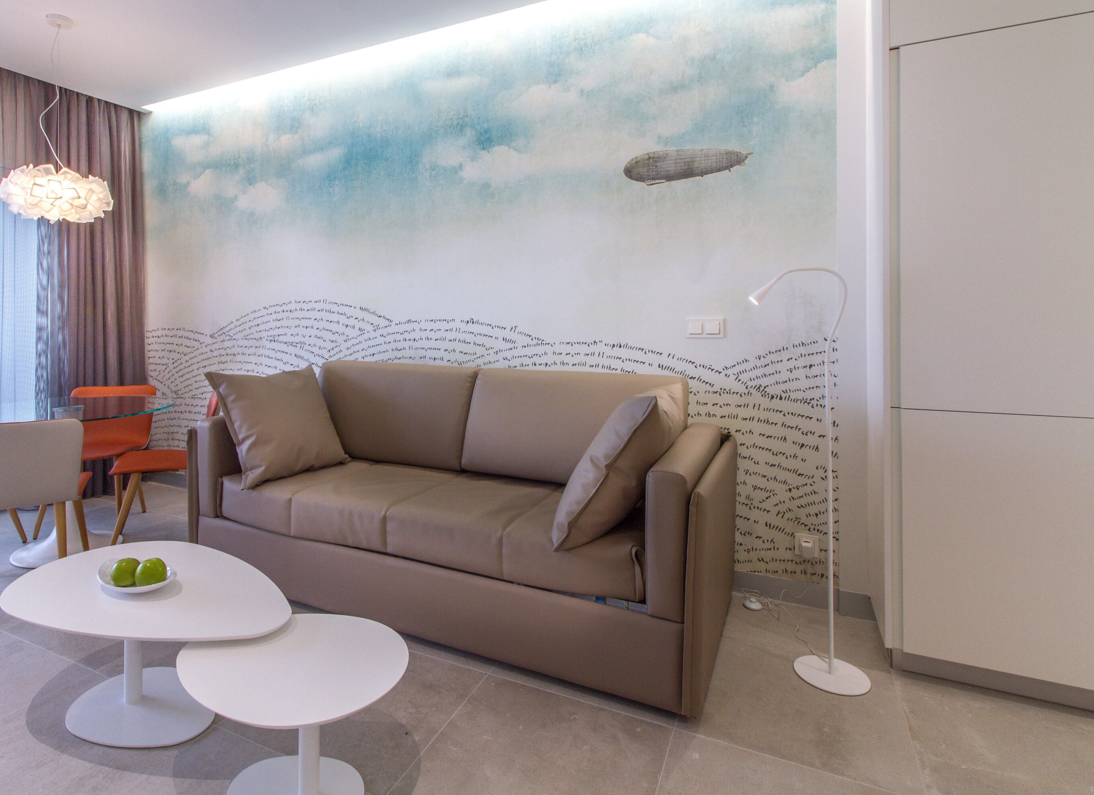 A modern living room with a beige sofa, two white round tables, and a wall mural depicting clouds and a zeppelin.