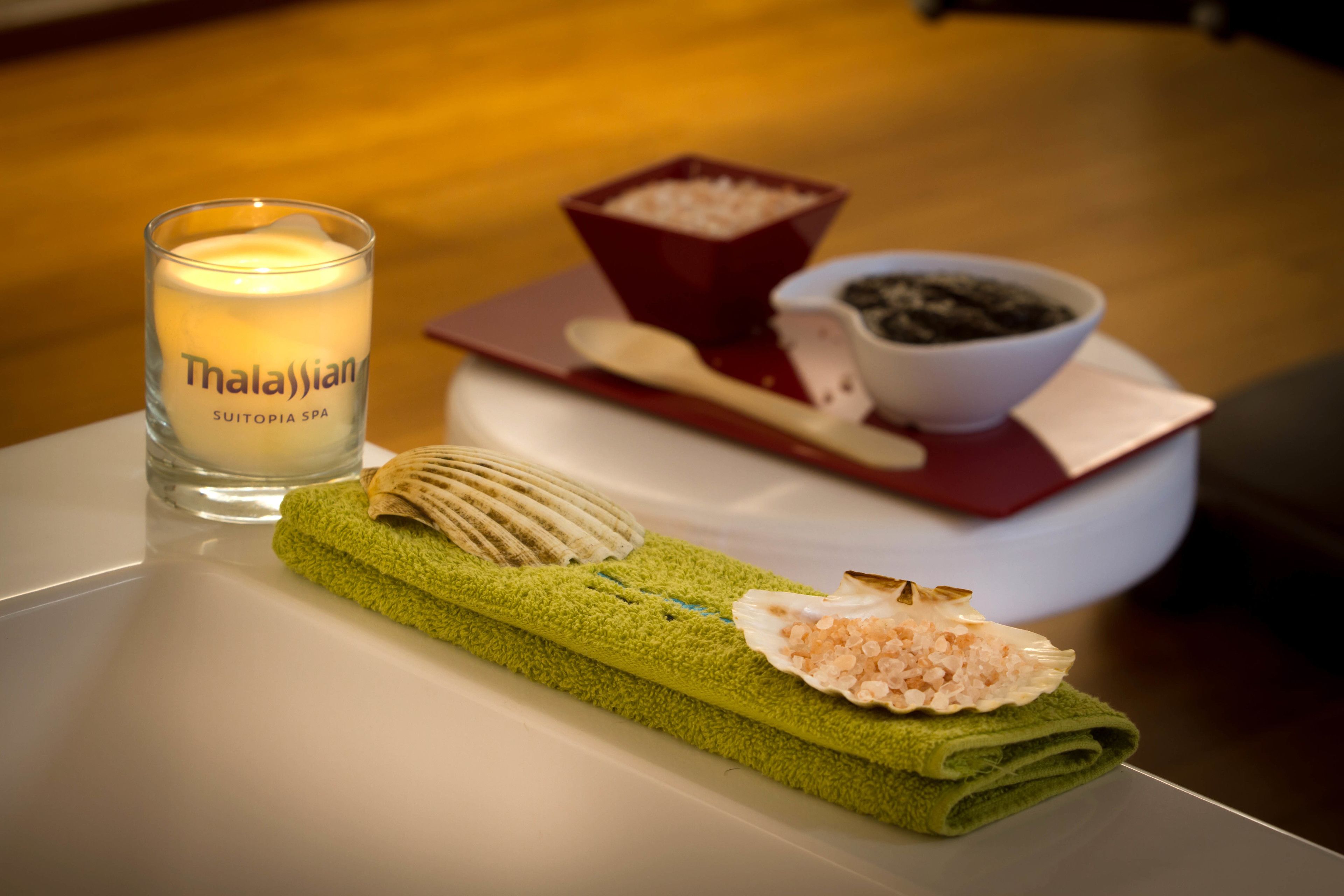 A candle labeled "Thalassian Suitopia Spa" on a table with a green towel, a shell, a bowl of black pebbles, and a spoon.