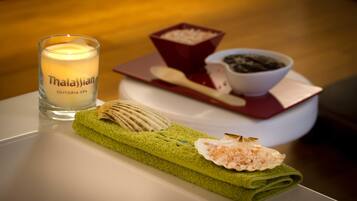 A candle labeled "Thalassian Suitopia Spa" on a table with a green towel, a shell, a bowl of black pebbles, and a spoon.