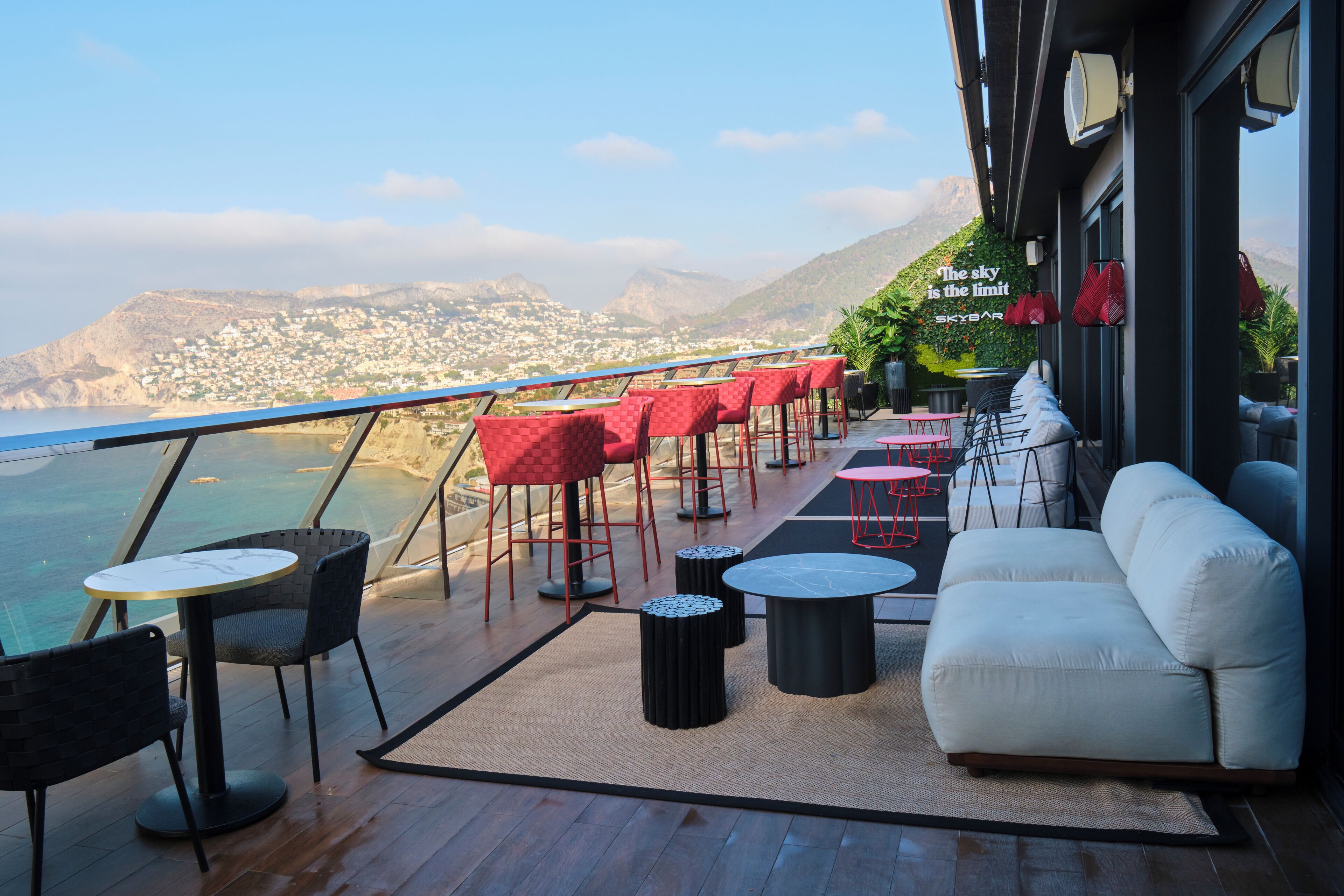 A terrace with a view of the sea and mountains, featuring red and black furniture, a white sofa, and a SkyBar sign.