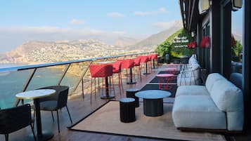 A terrace with a view of the sea and mountains, featuring red and black furniture, a white sofa, and a SkyBar sign.