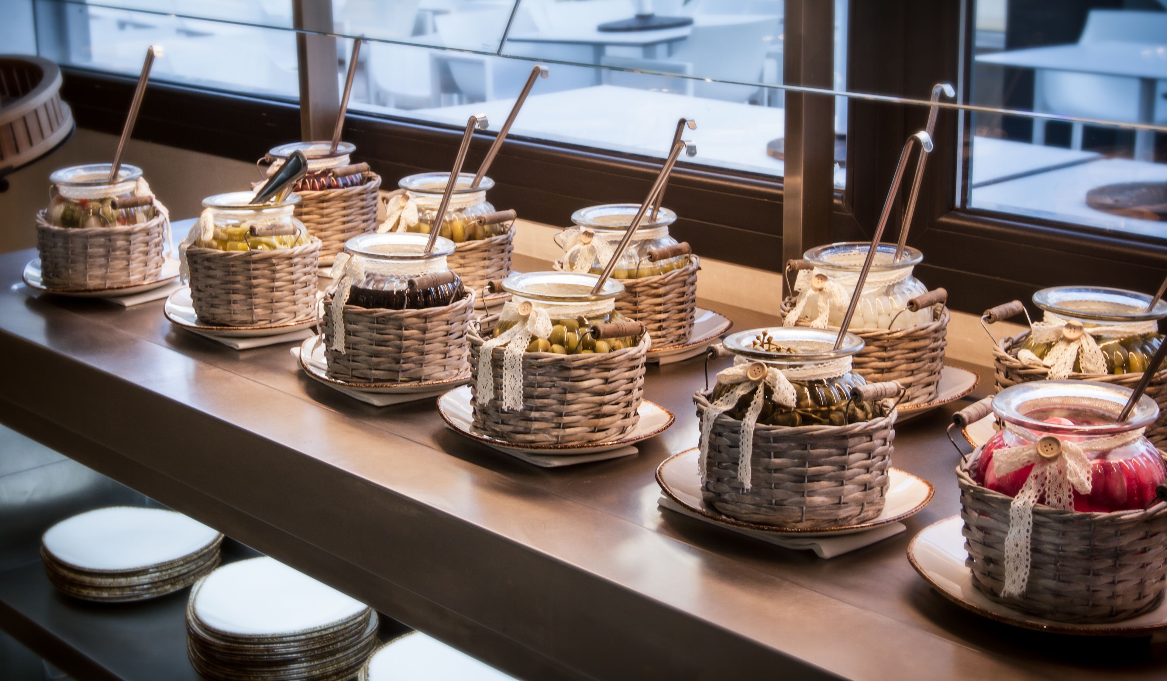 A buffet setup with various food items in wicker baskets and glass jars, arranged on a counter with plates underneath.
