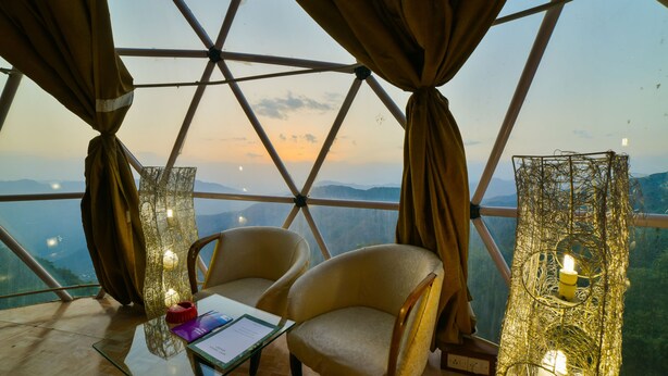 A room with a large window, two chairs, a table with a menu, and a decorative light fixture. - Mint Tarika Resort (Kandaghat)