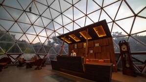 A reception area with a wooden desk, a clock, and a view of the outdoors through a large glass dome structure. - Mint Tarika Resort (Kandaghat)