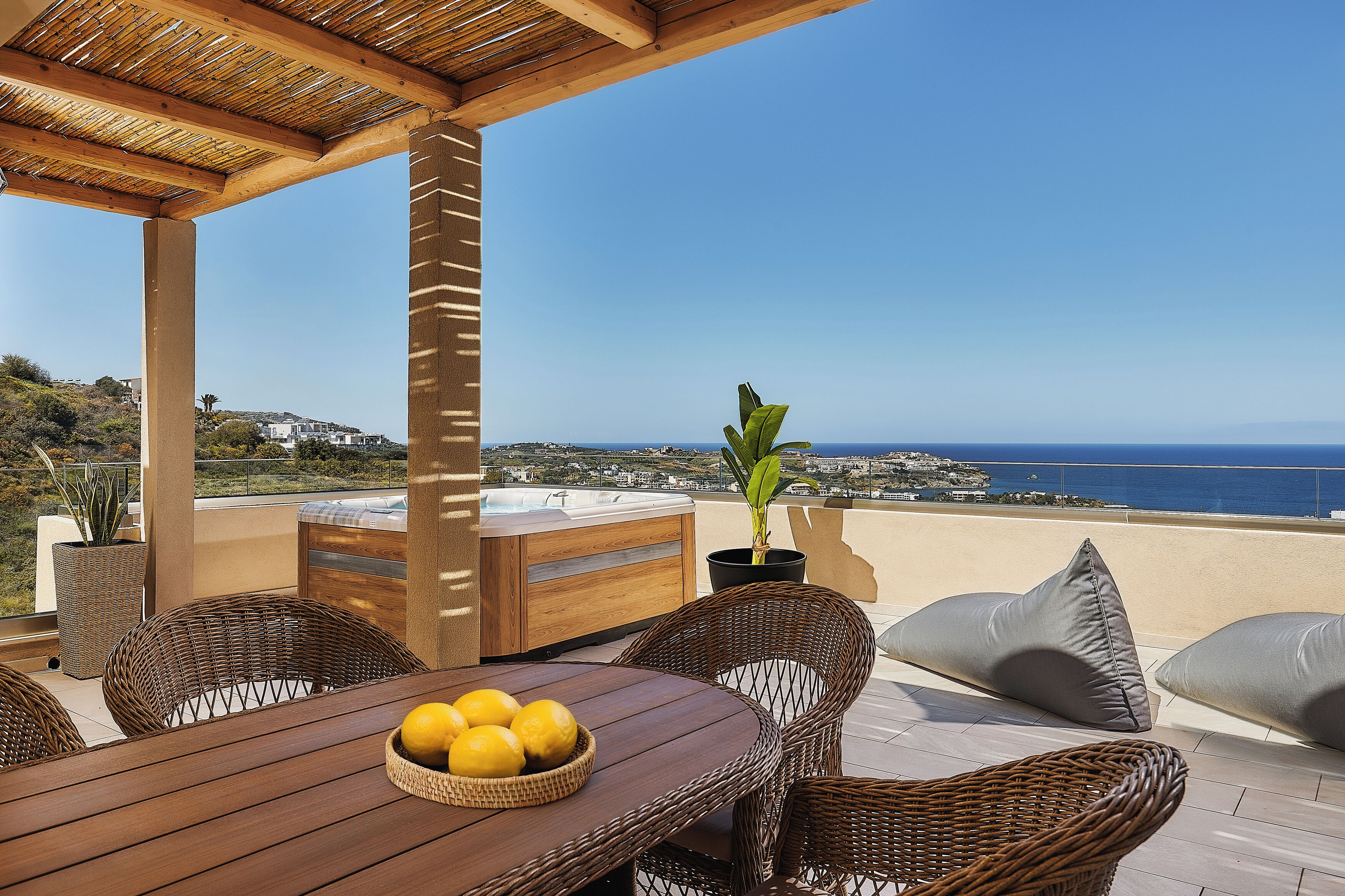 Una mesa de madera con un bol de limones, sillas de mimbre, una bañera de hidromasaje y vistas al mar.