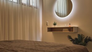 A bedroom with a bed, a round mirror, a shelf with a plant, and a window with curtains. - OZEA - Elevated Living (Malevizi)