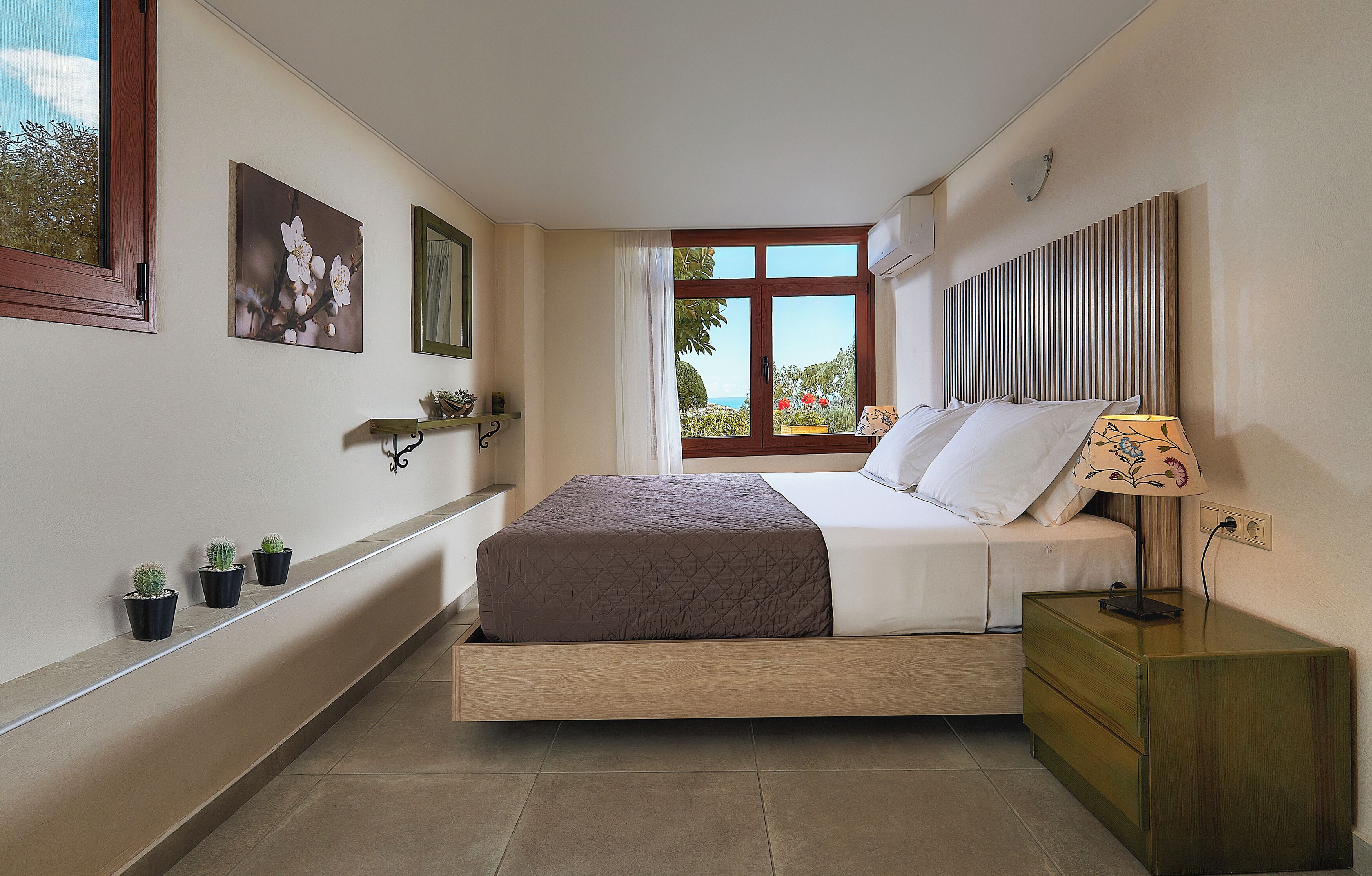 A bedroom with a bed, bedside table, lamp, potted plants, and a window with a view.