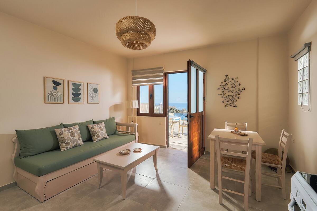 A living room with a sofa, coffee table, and dining table with chairs. There is a window with a view of the sea.