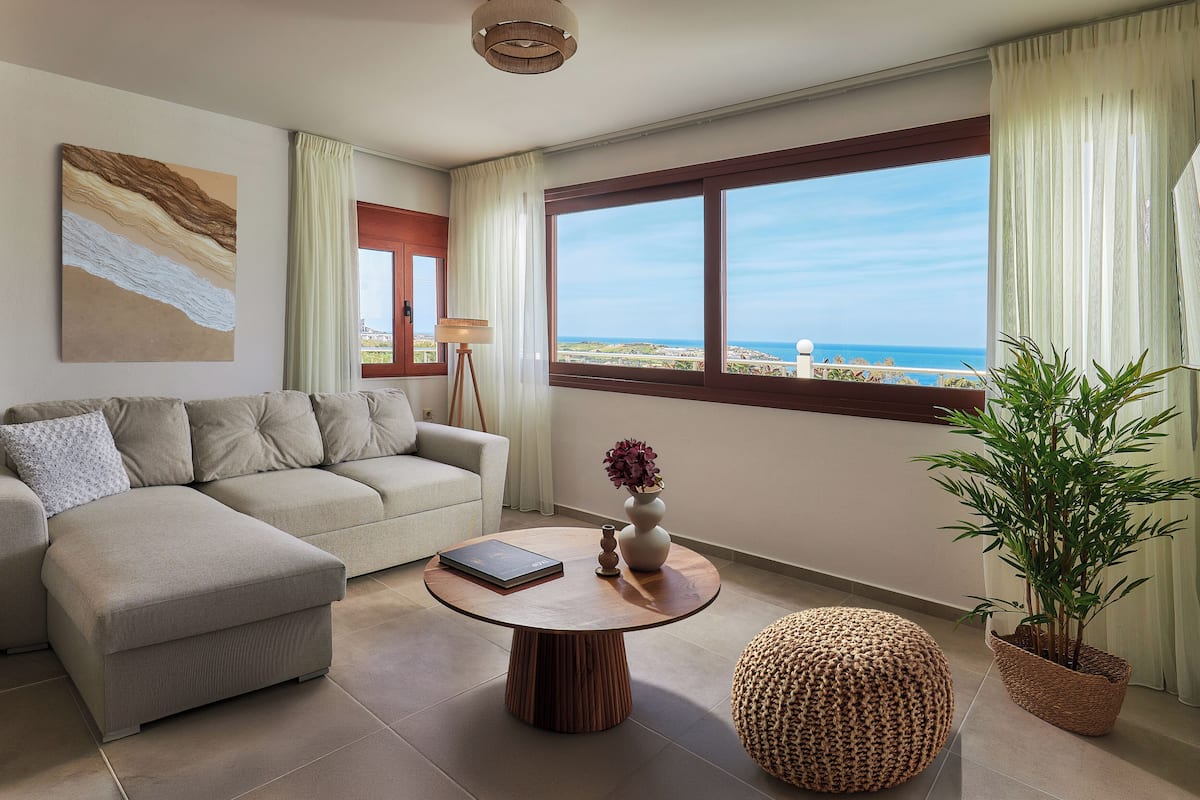 A modern living room with a sofa, a round coffee table, a potted plant, and a view of the ocean.