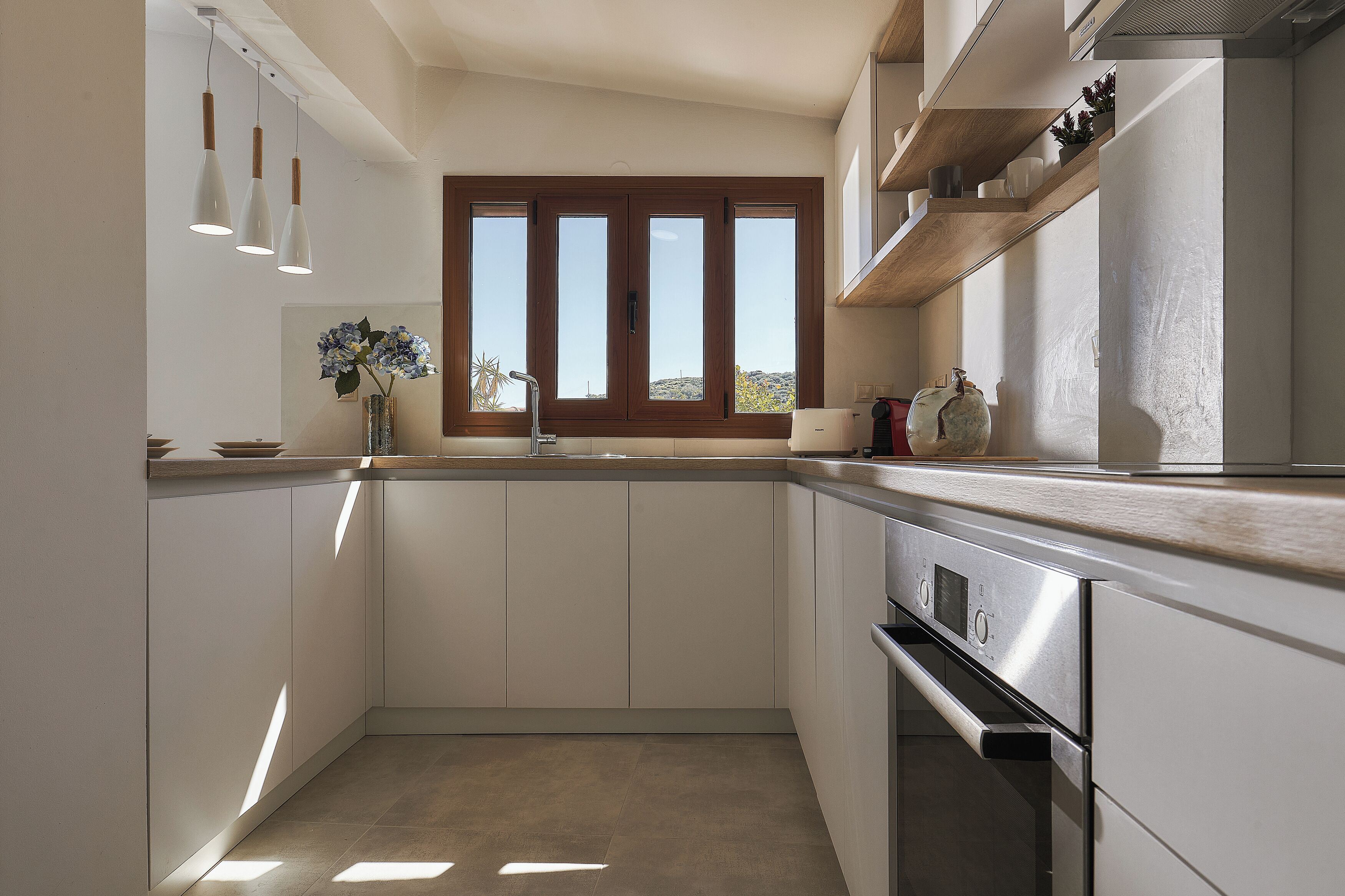 Una cocina con armarios blancos, horno de acero inoxidable, ventana con persianas e iluminación colgante.