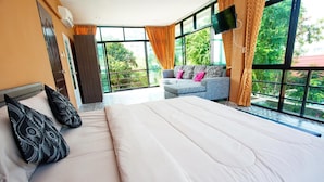 Habitación de hotel con una cama grande, un sofá con cojines, un televisor y ventanas amplias con vistas a zonas verdes.