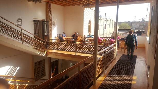 A multi-level building with wooden railings and a staircase. There are tables and chairs on the upper level, and a person is walking on the lower level. - Islambek Hotel & Travel (Khiva)