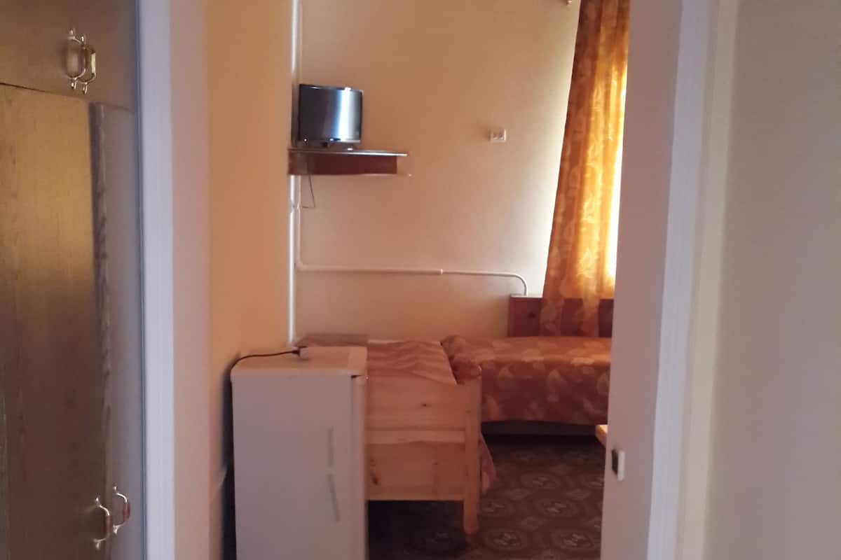 Ein schmaler Gang führt zu einem Schlafzimmer mit einem Bett, einem kleinen Tisch und einem Heizgerät.