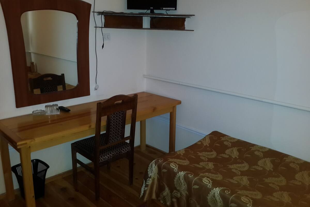 Ein Zimmer mit einem Holztisch, einem Stuhl, einem Bett mit gemustertem Überwurf und einem an der Wand befestigten Fernseher sowie einem Spiegel über dem Tisch.