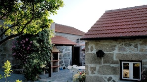 Un bâtiment en pierre avec un toit recouvert de tuiles rouges, un petit jardin parsemé de fleurs roses et une cour dallée.