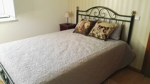 A neatly made bed with a patterned bedspread and two decorative pillows, a bedside table with a lamp, and a window with blinds. - Quinta de Vodra (Seia)