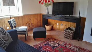 Una sala de estar con sofá, reposapiés y televisor sobre una pared de madera. Hay dos cestas y un jarrón decorativo con flores.