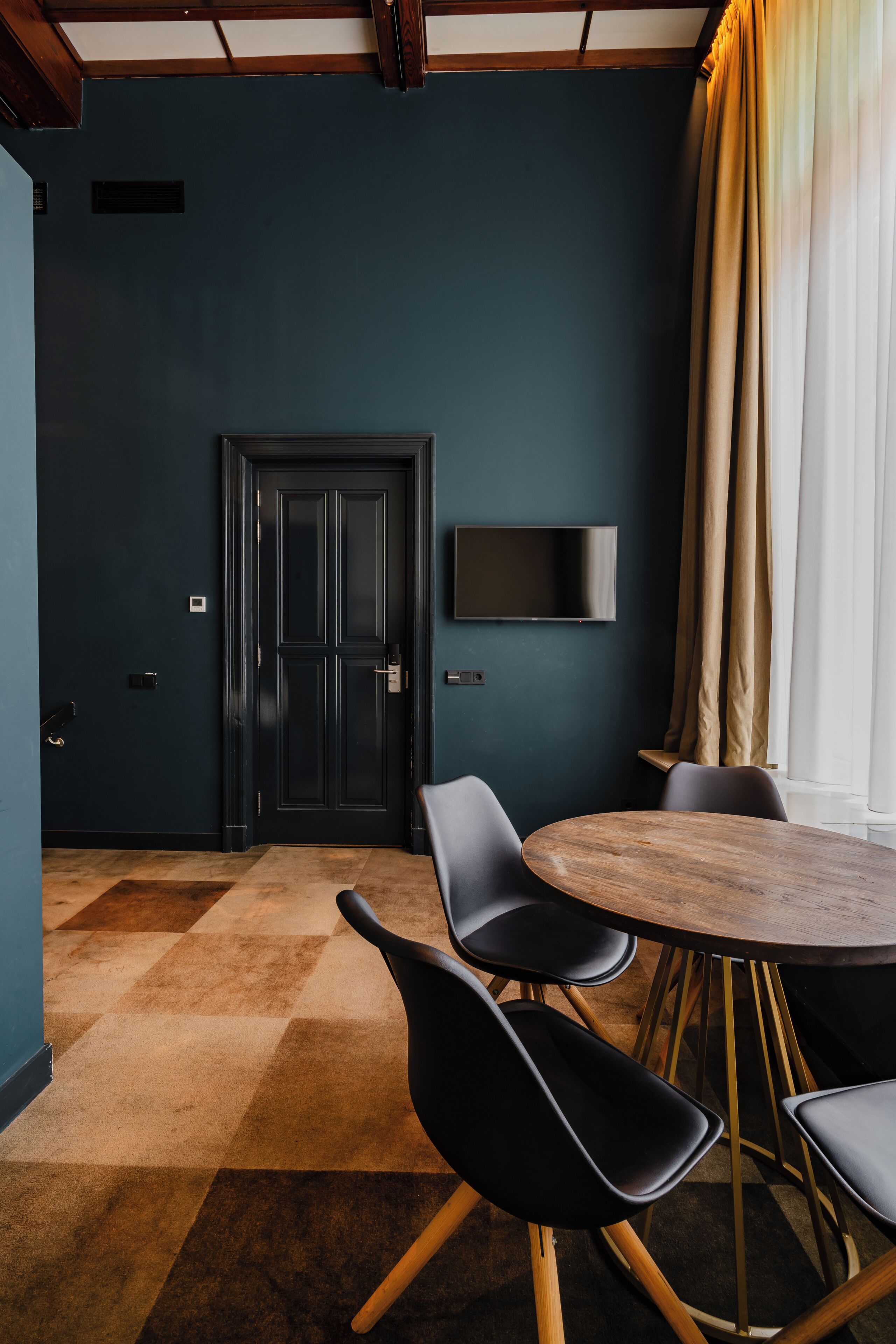 Une pièce avec un mur bleu foncé, une porte en bois, un téléviseur à écran plat fixé au-dessus, et une table ronde entourée de quatre chaises noires.