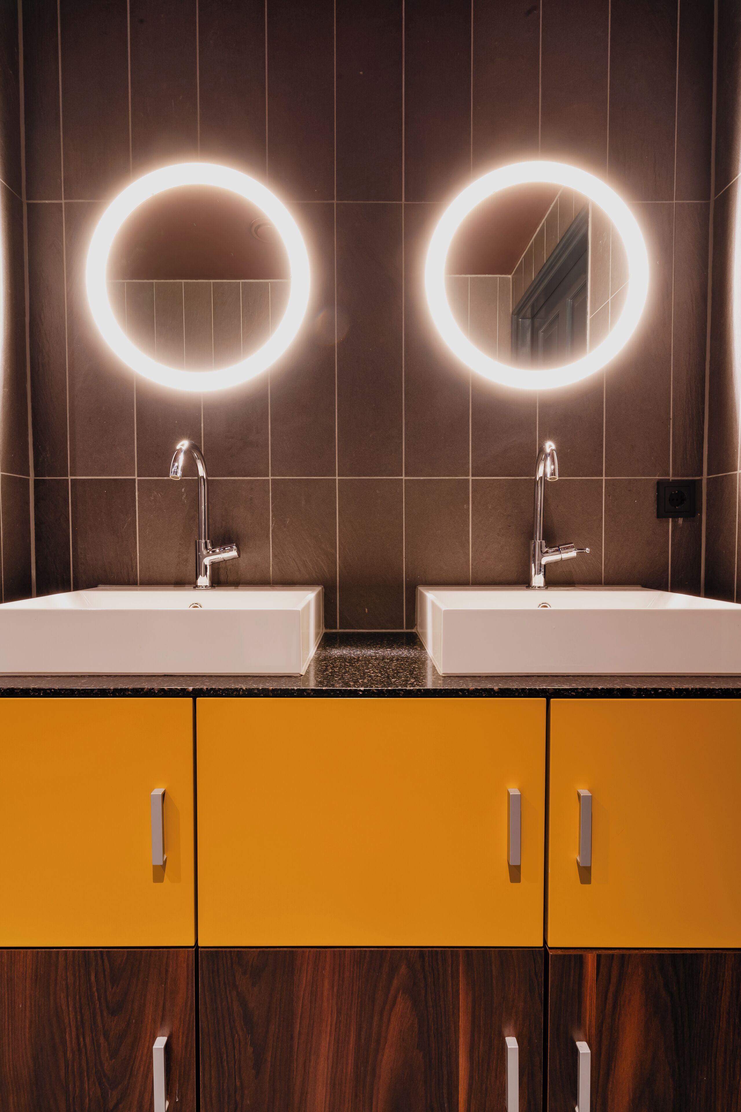 Een badkamer met twee wastafels, elk voorzien van een spiegel en verlicht met een ringlamp. De wanden zijn betegeld, en er zijn houten kastjes onder de wastafels.