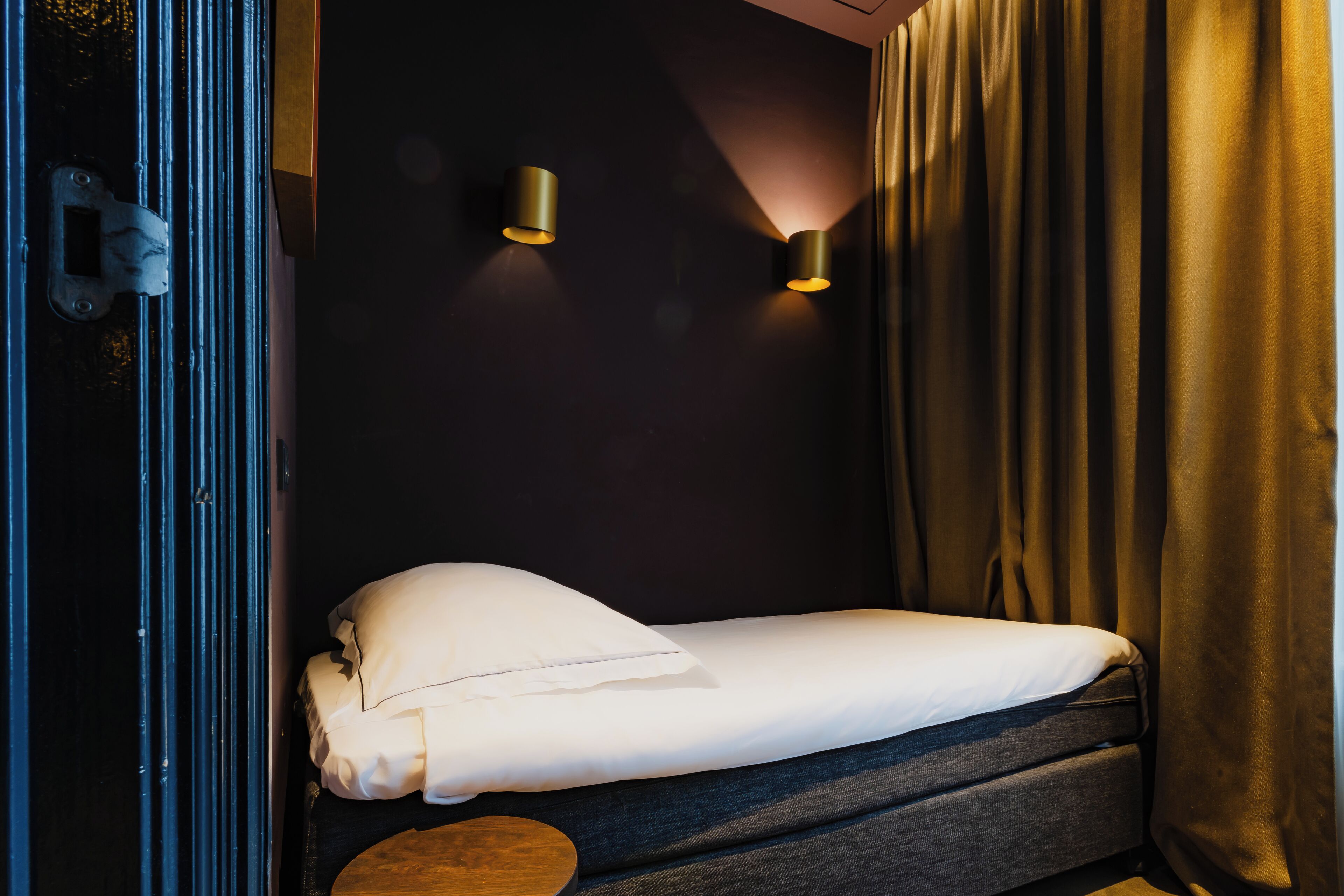 Un lit simple avec un oreiller et une couverture blancs, une petite table ronde et deux lampes fixées au mur.