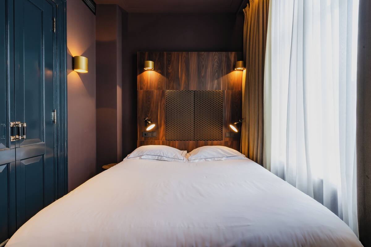A neatly made bed in a hotel room with a wooden headboard, two wall sconces, and a large window with curtains.
