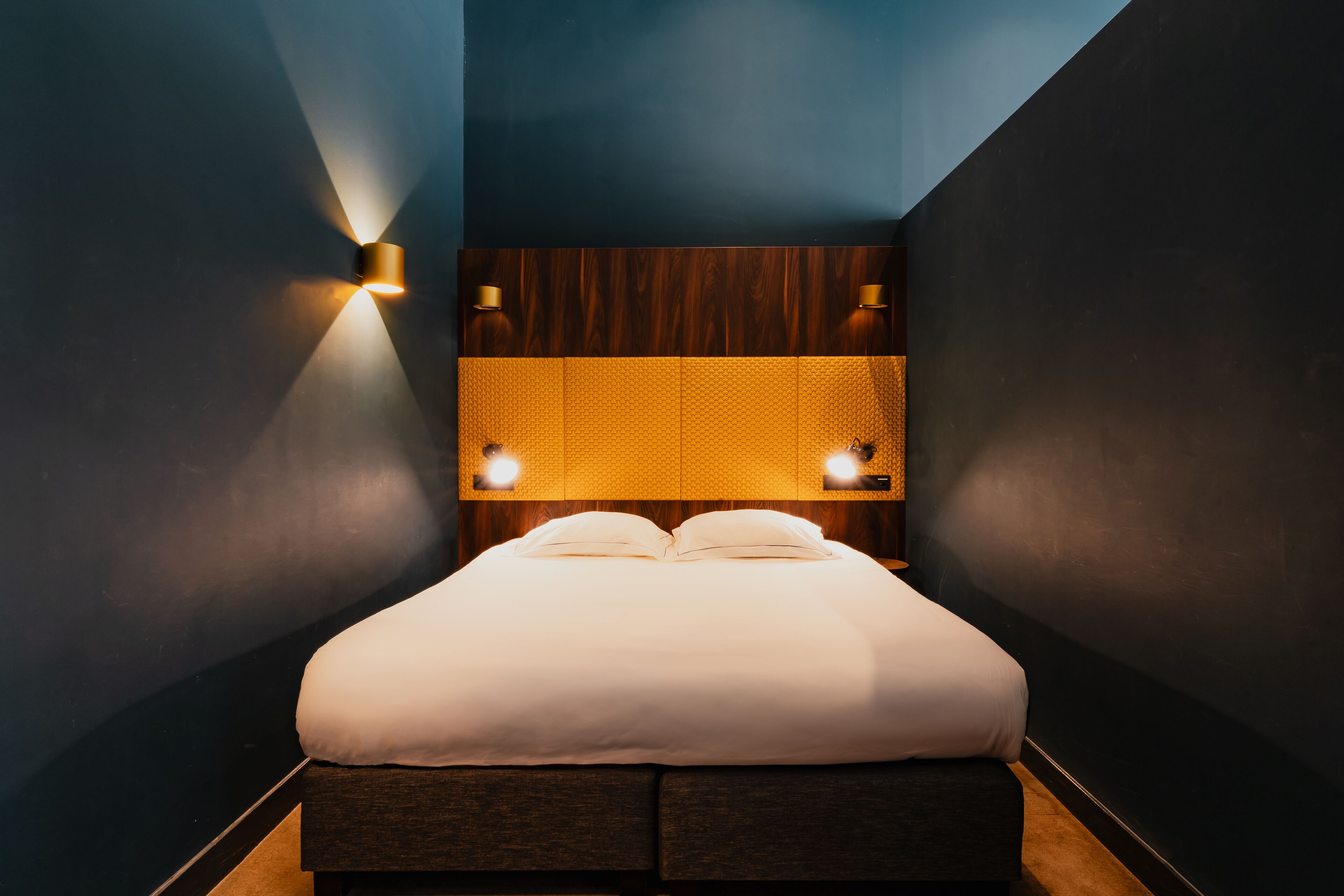 Ein modernes Hotelzimmer mit einer dunkelblauen Akzentwand, einem Holzkopfteil mit strukturierter Paneeleinlage und zwei Wandleuchten.