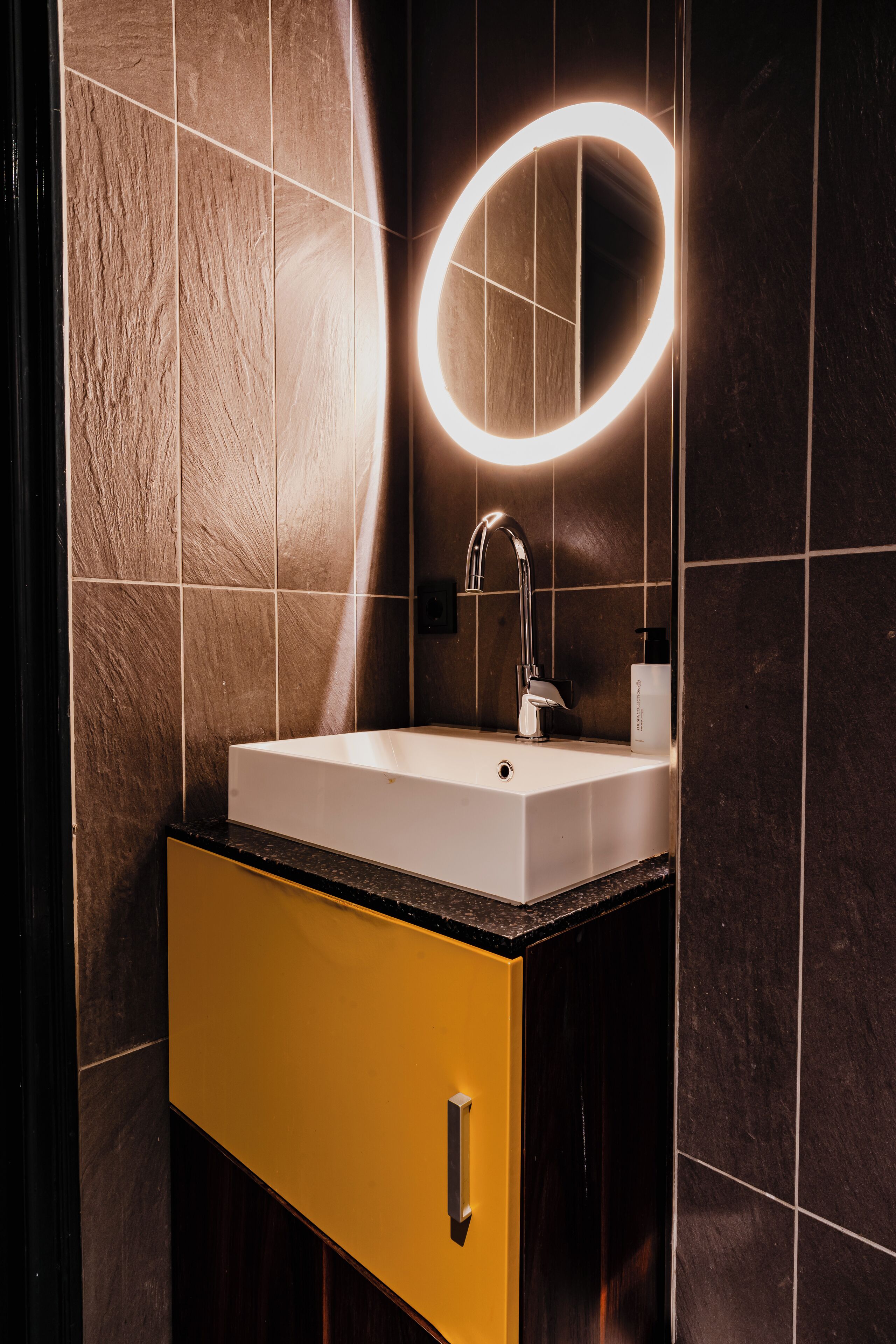 A bathroom with a white sink, a mirror with a glowing light, and dark brown tiles.