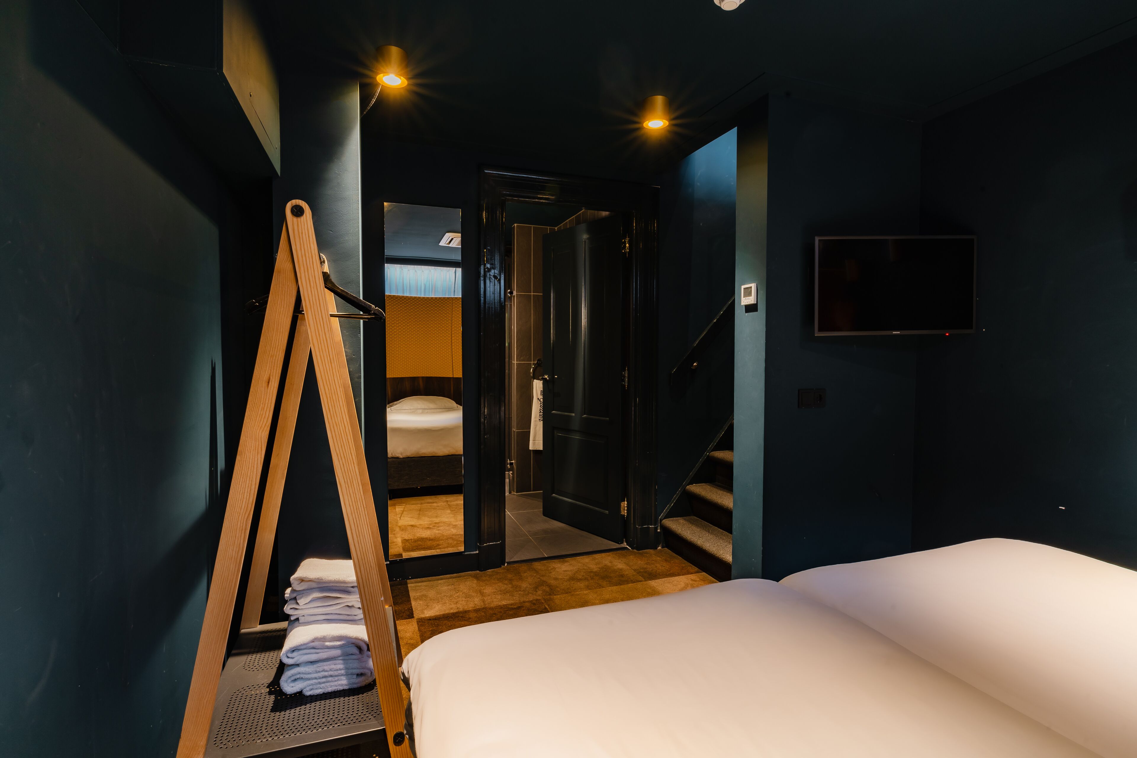 A hotel room with a bed, a wooden ladder, a small table with towels, a TV, and a staircase leading to a bathroom.
