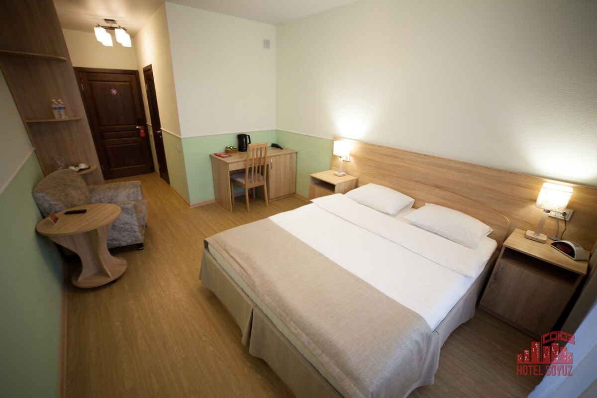 A hotel room with a bed, bedside tables, a desk with a chair, a small table, and a wardrobe.