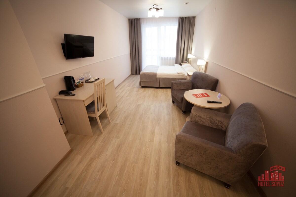 A hotel room with a bed, a desk, a chair, a sofa, a TV, and a lamp.