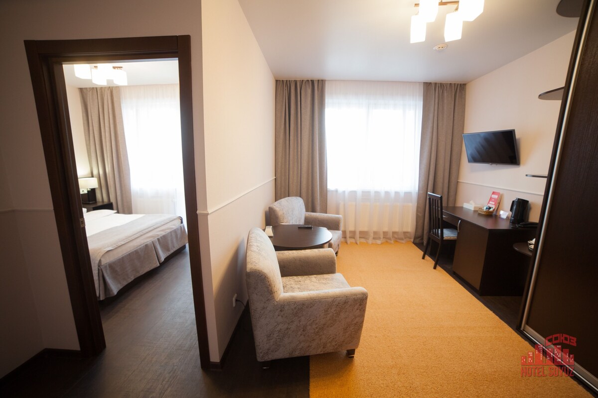A hotel room with a bed, a sofa, a desk, a TV, and a window with curtains.