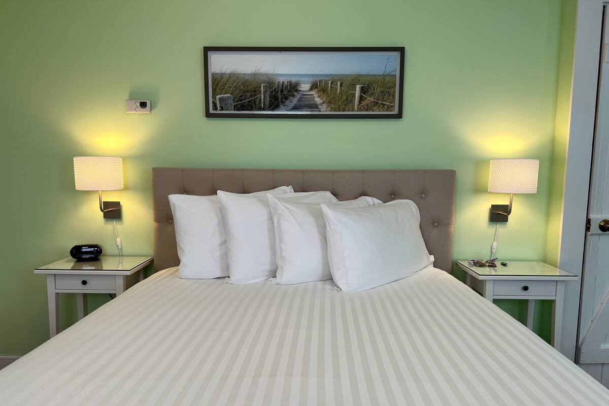 Cama bem arrumada com colcha listrada, duas mesas de cabeceira com luminárias, um quadro emoldurado sobre a cama e uma porta à direita.