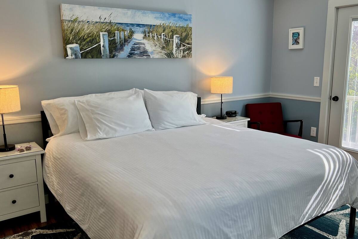 Cama bem arrumada com lençóis brancos, duas luminárias de cabeceira, uma mesinha de cabeceira e um quadro com uma paisagem costeira na parede.