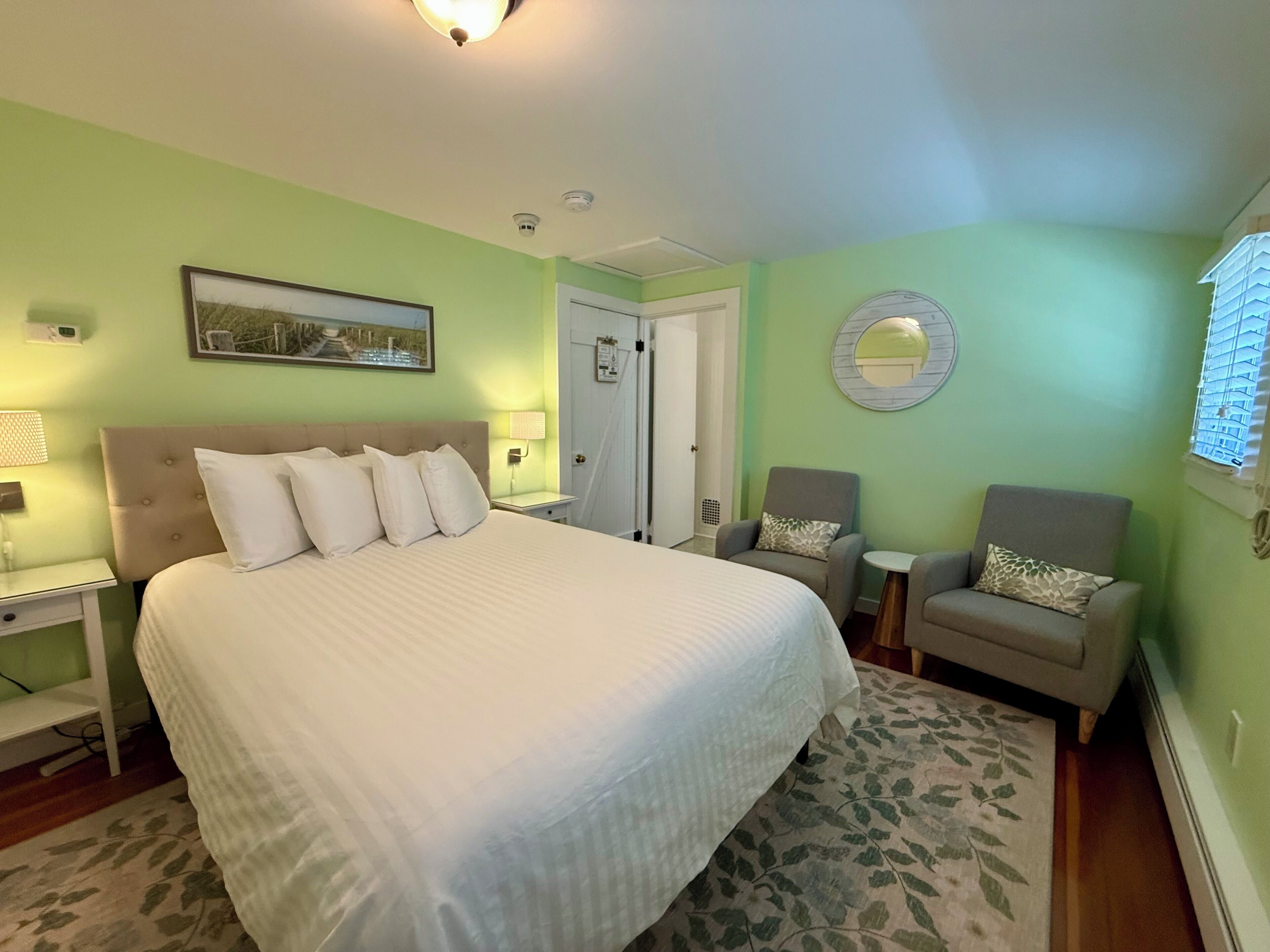 A bedroom with a large bed, two armchairs, a small table, a mirror, and a door.