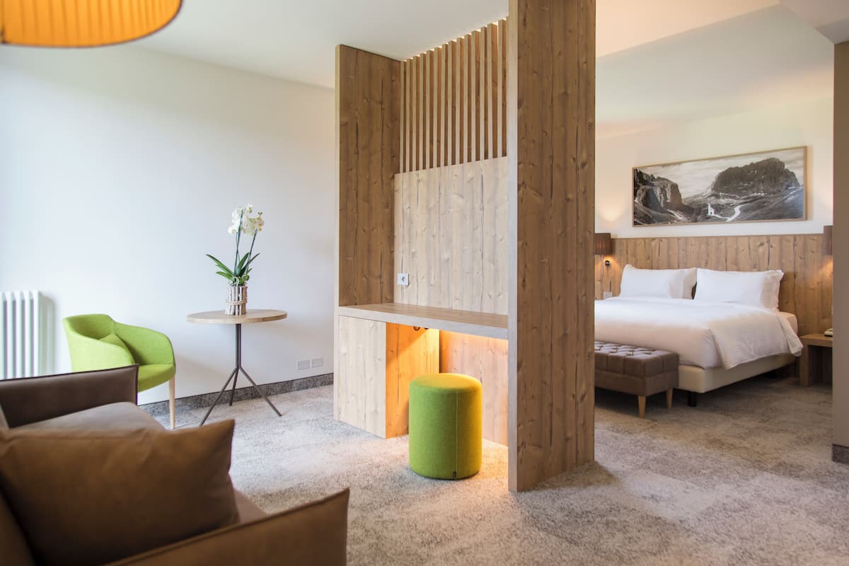 A modern hotel room with a bed, a green chair, a small table with a plant, and a wooden partition.