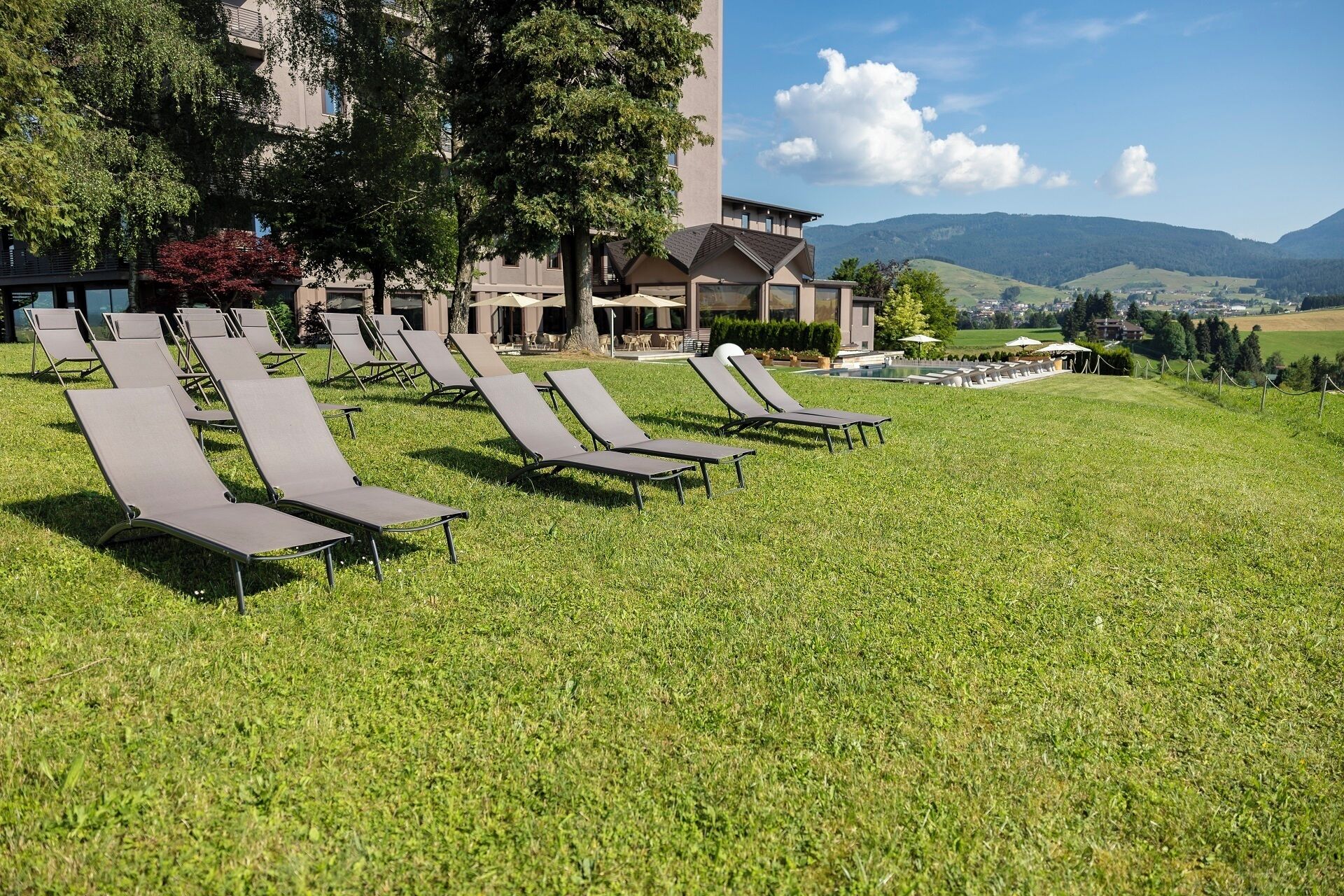 A hotel with outdoor lounge chairs, a pool, and a view of mountains.