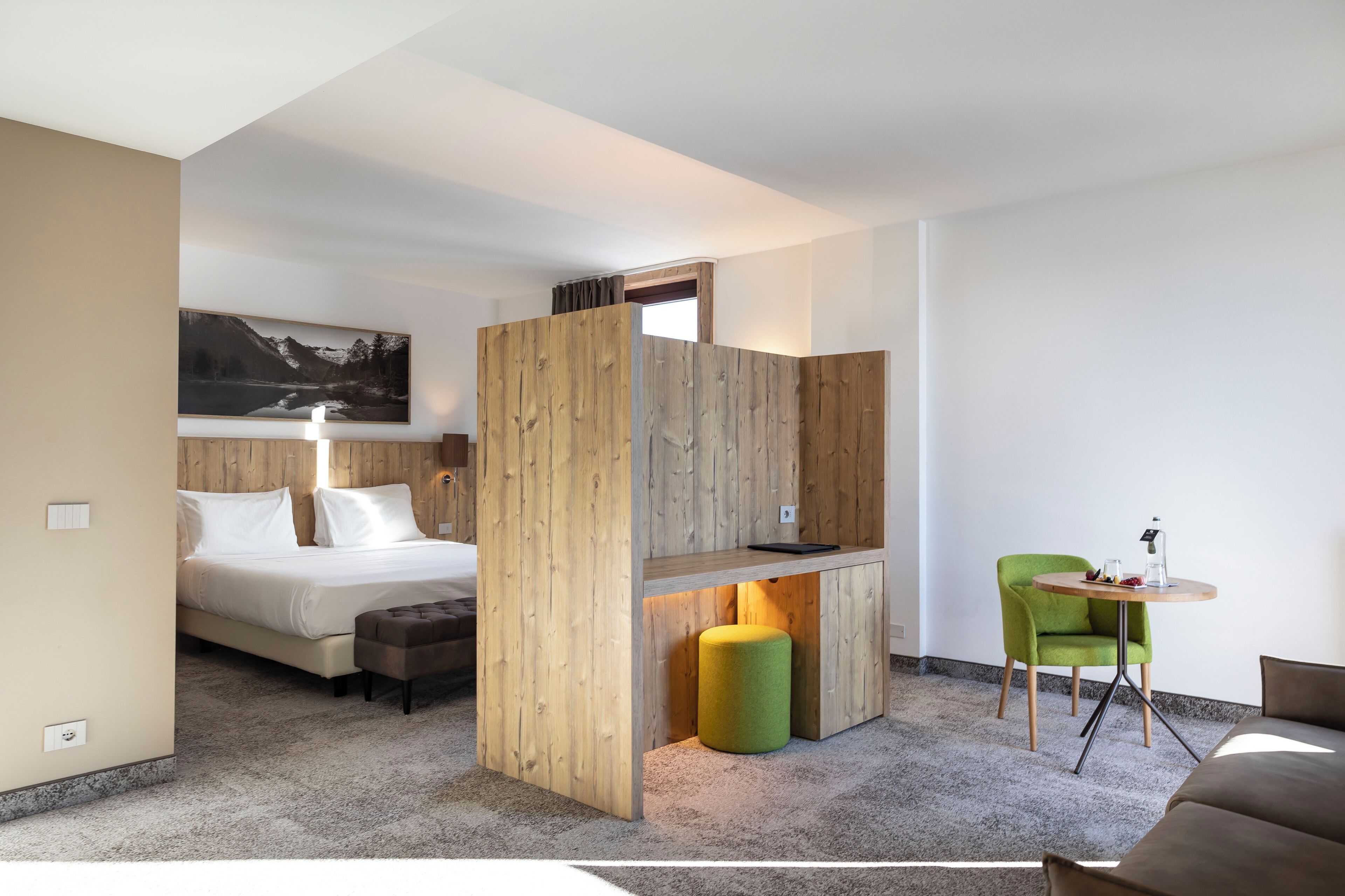 A modern hotel room with a bed, a wooden partition, a green chair, a small table, and a painting on the wall.