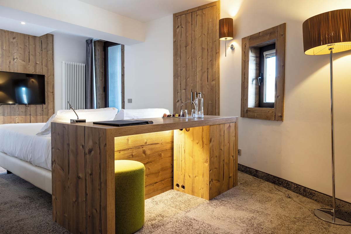 A modern hotel room with a wooden accent wall, a bed, a small desk with a lamp, and a TV mounted on the wall.