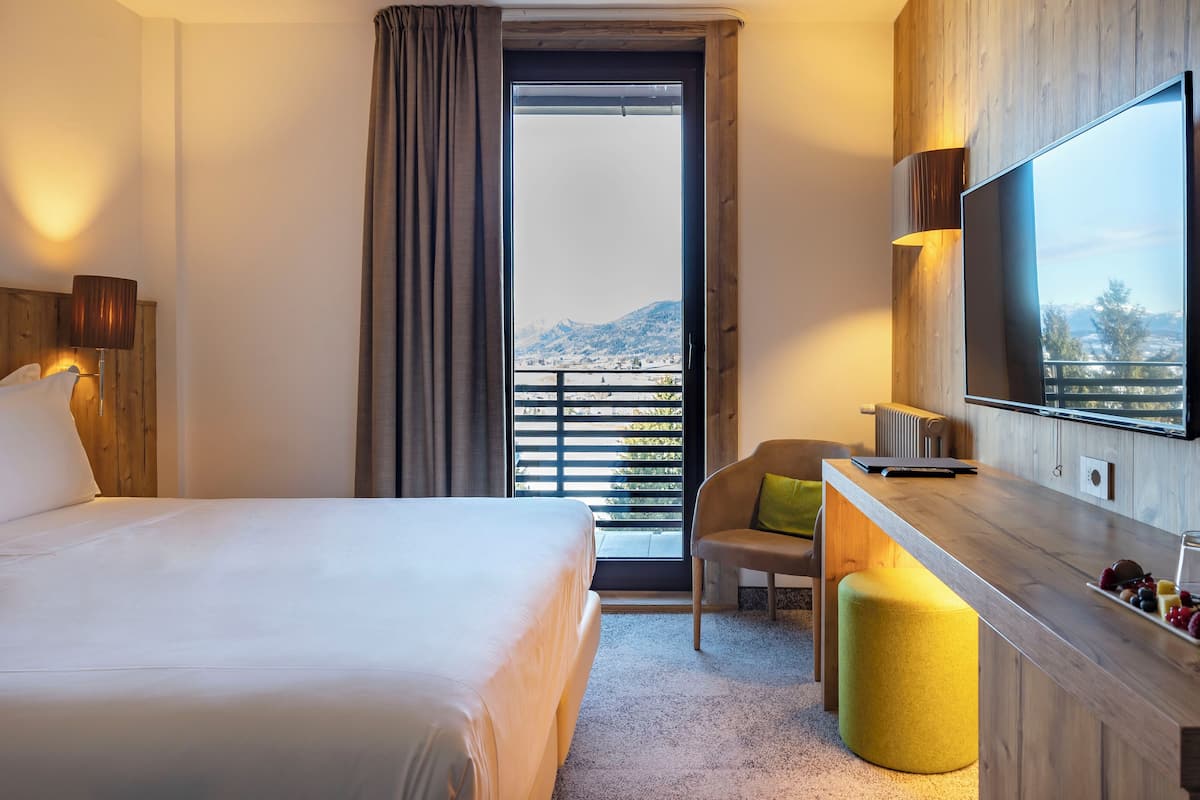 A hotel room with a bed, a chair, a small table with a glass of water, a TV, and a view of mountains through a large window.
