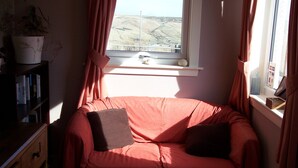 A cozy room with a red sofa, a bookshelf, and a window with a view of a hilly landscape. - Strathy Inn (Thurso)