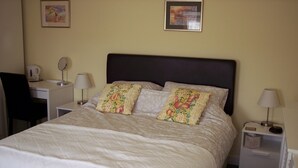 A neatly made bed with floral pillows, a bedside table with a lamp, a chair, and a small nightstand with a lamp and a tissue box. - Strathy Inn (Thurso)