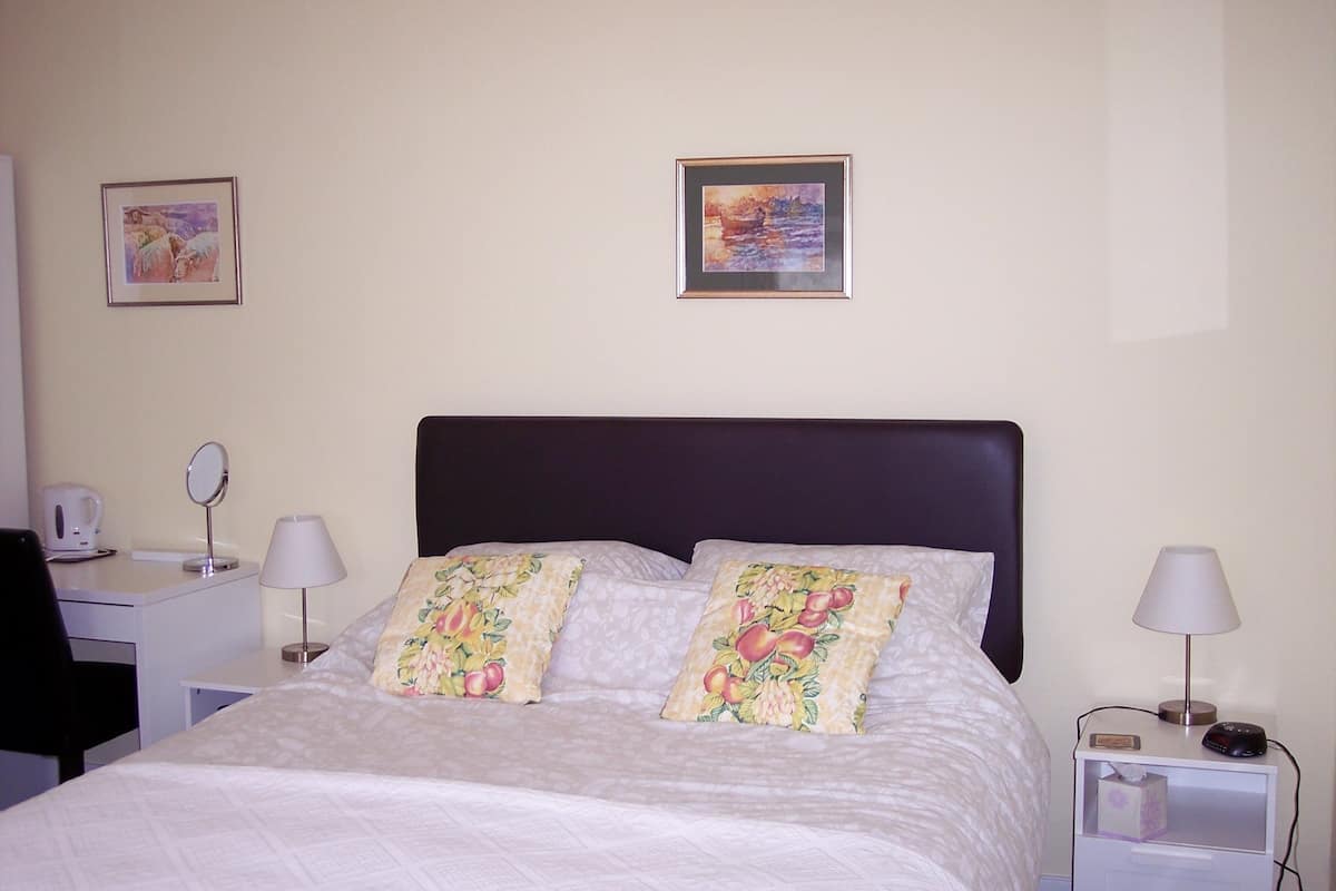A neatly made bed with floral pillows, a bedside table with a lamp, a nightstand with a lamp, and two framed pictures on the wall.