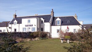 A two-story building with a sign reading "Strathy Inn." It features a stone wall, a grassy lawn, and some shrubs. - Strathy Inn (Thurso)