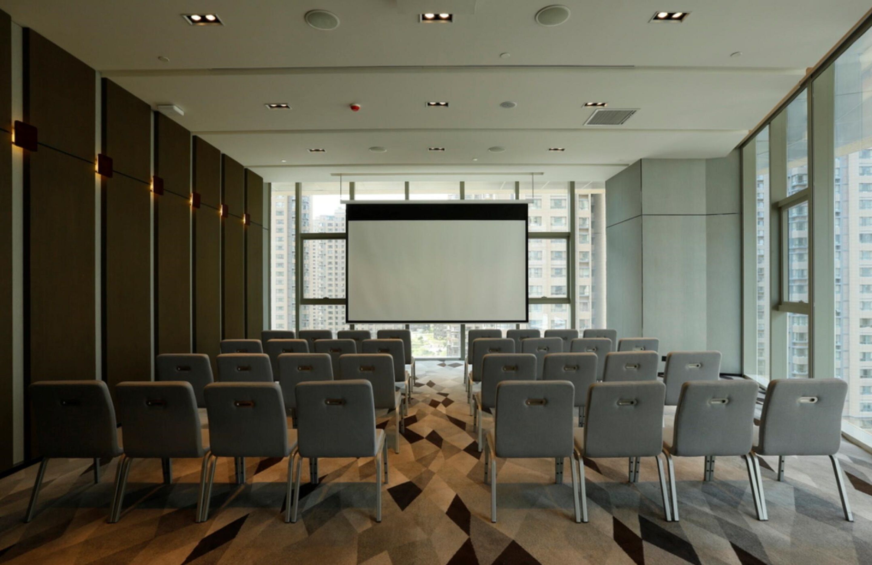 Une salle de conférence avec des rangées de chaises disposées face à un grand écran, de grandes fenêtres et un tapis à motifs.