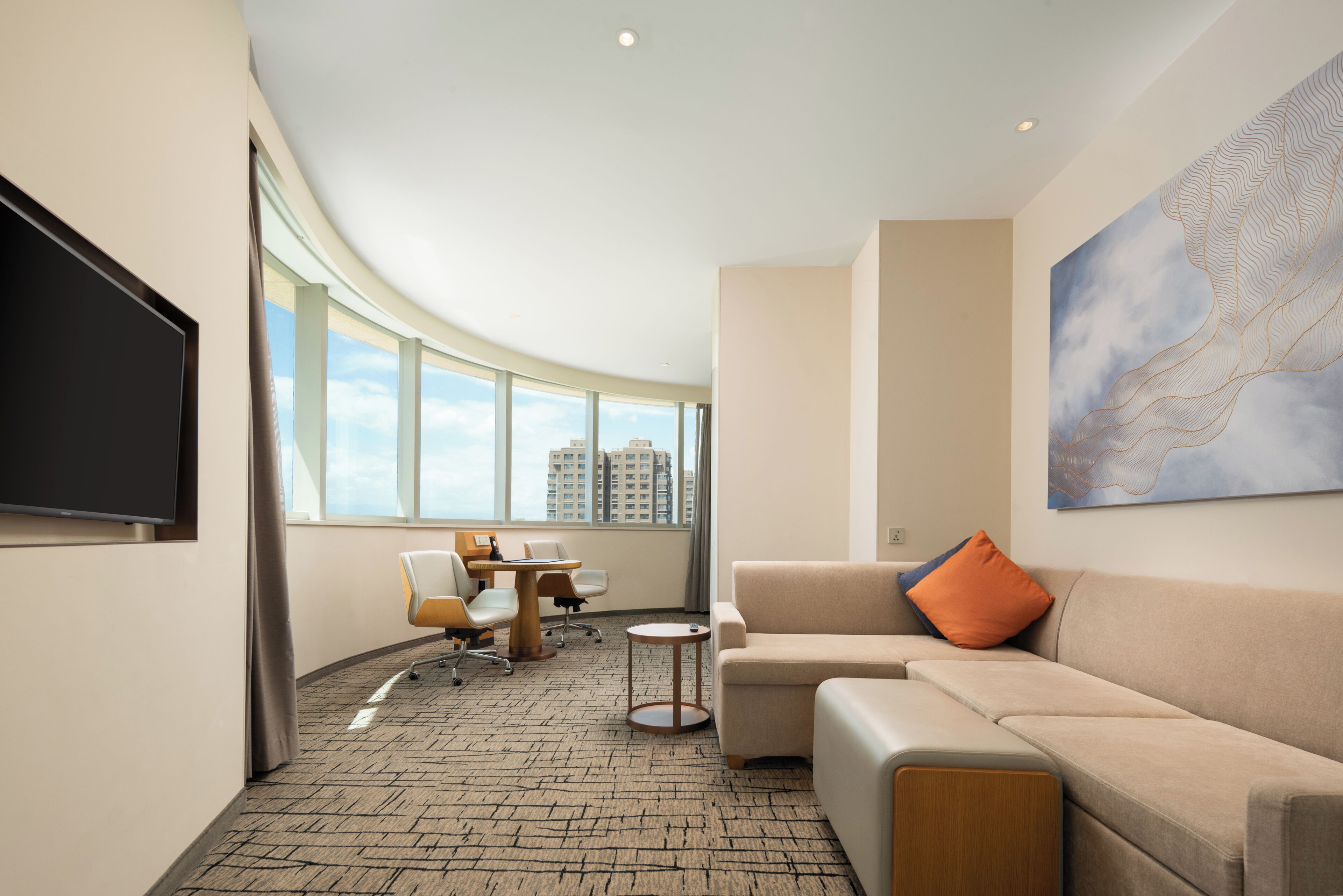 A modern hotel room with a curved ceiling, a flat-screen TV, a sofa, a coffee table, a desk with chairs, and a large window offering a city view.
