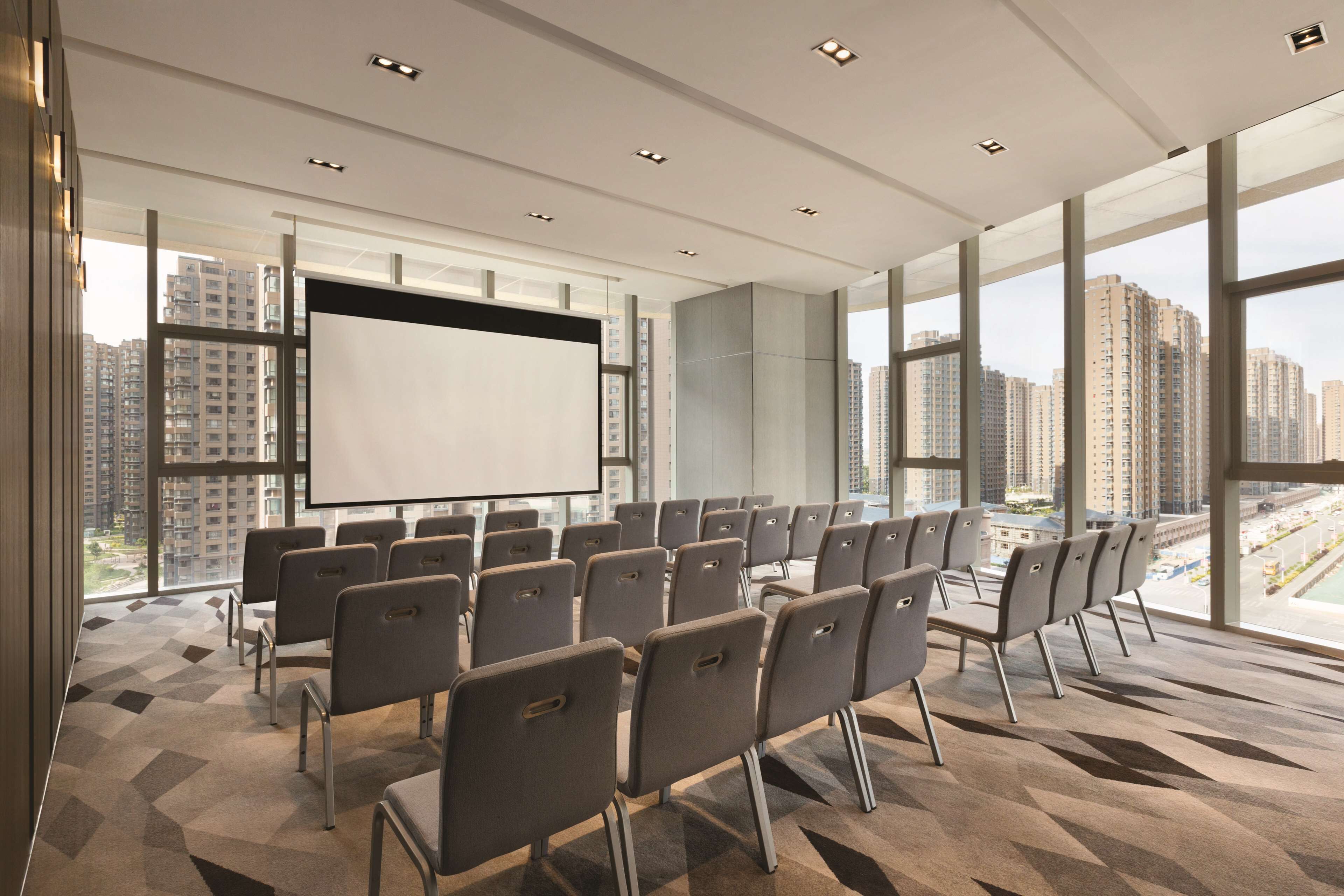 A conference room with a large screen, rows of chairs, and large windows offering a city view.