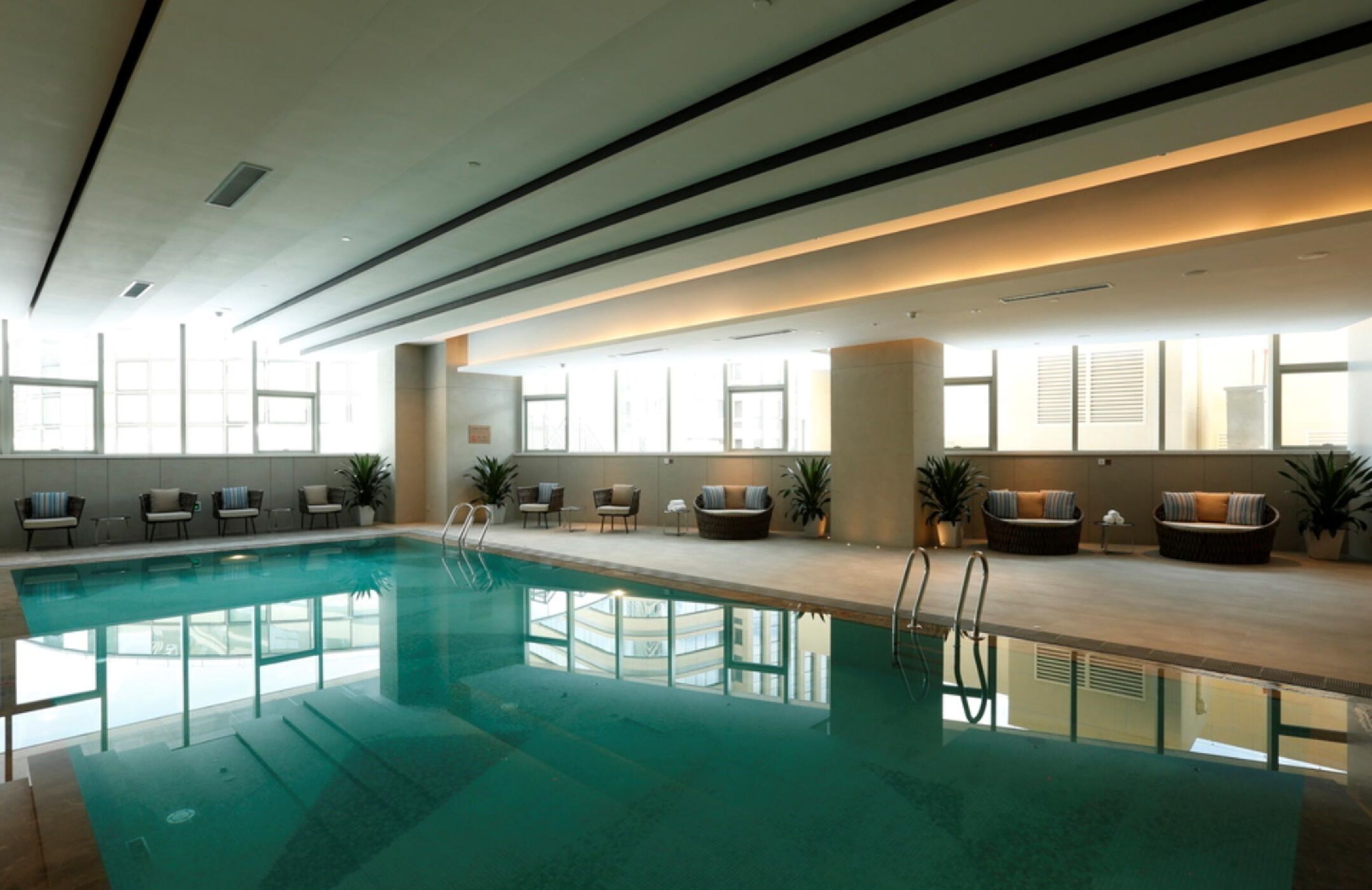 Ein Hallenbad mit klarem Wasser, umgeben von Liegestühlen und Sofas, großen Fenstern und einem modernen Deckenbau.