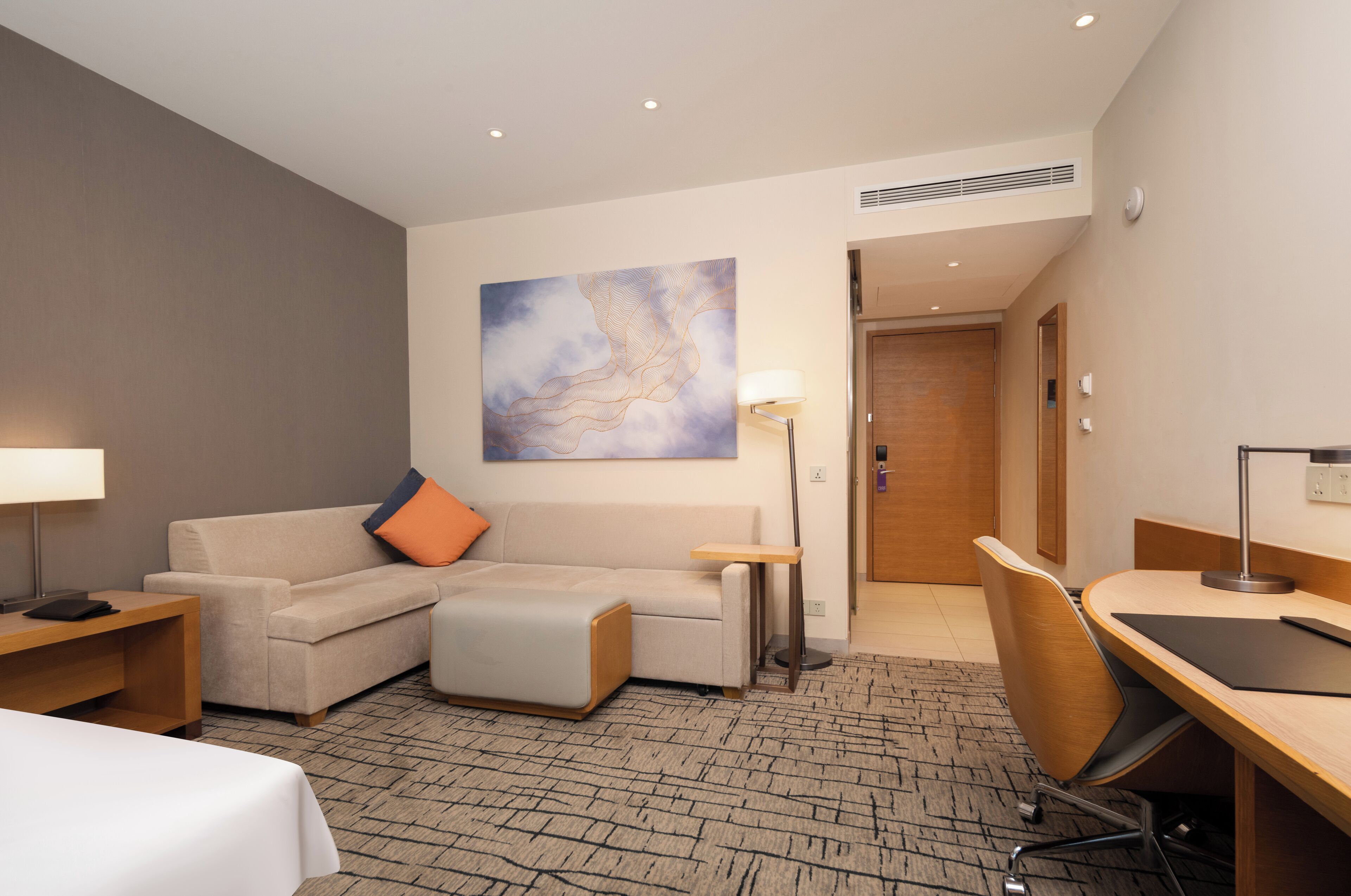 A hotel room with a sofa, a desk, a lamp, a painting on the wall, and a door leading to another room.