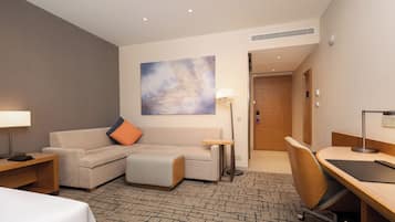 A hotel room with a sofa, a desk, a lamp, a painting on the wall, and a door leading to another room.
